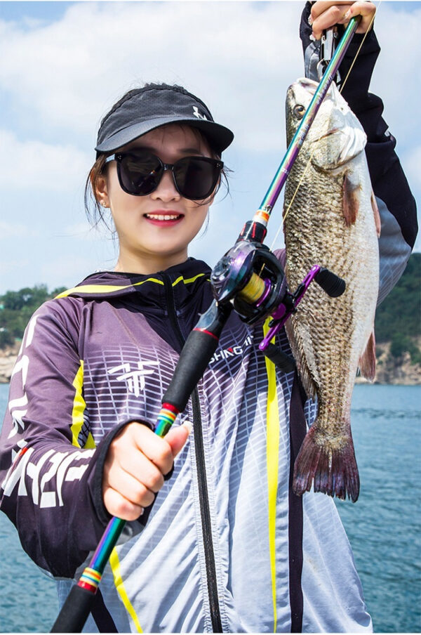 Joyful woman with stylish fishing gear proudly displays her vibrant catch by the water.