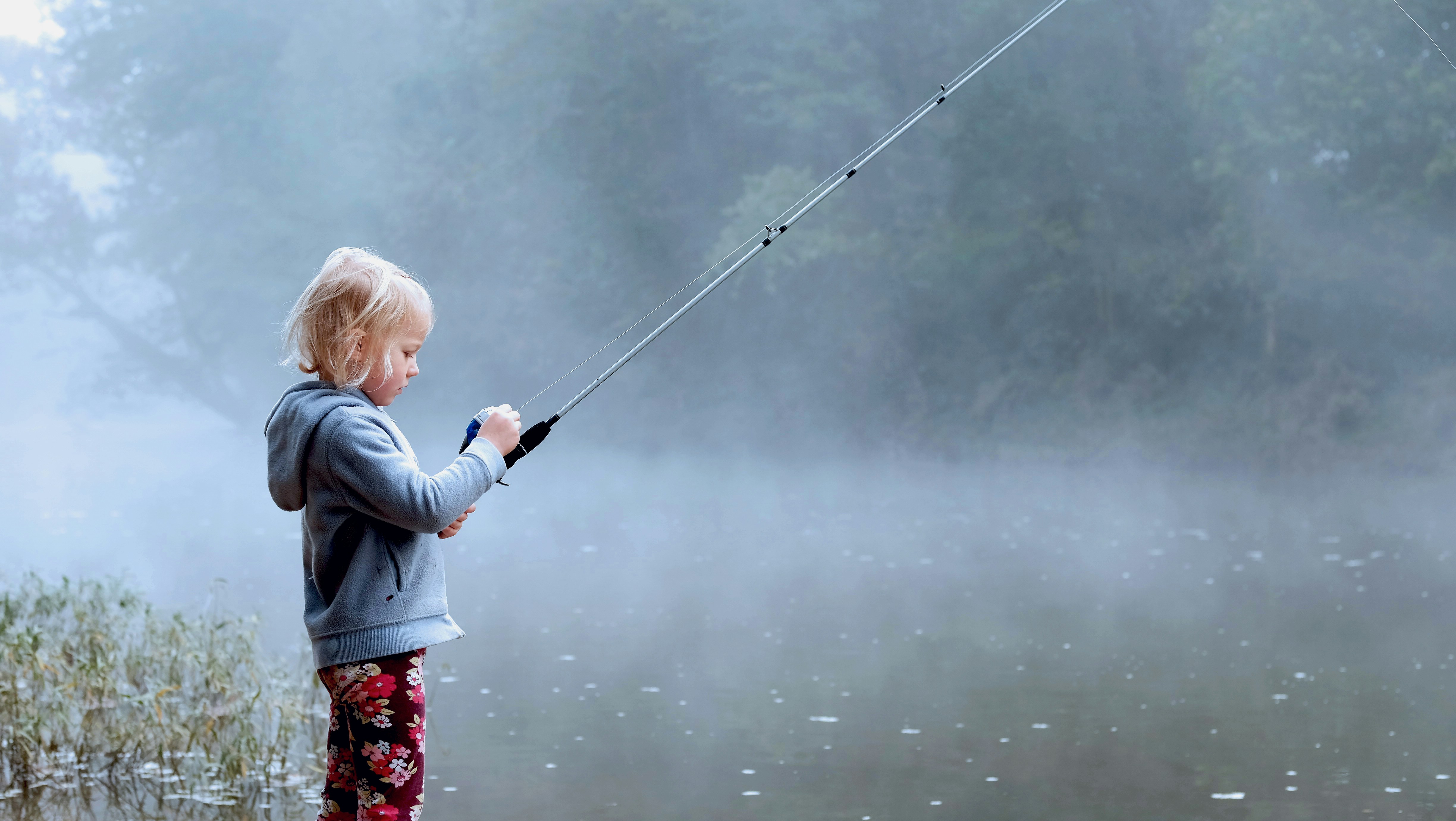 Young girl fishing peacefully by a misty lake in cozy attire, exploring natures beauty.