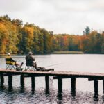 Kaksi kalastajaa rentoutuu syksyisellä laiturilla nauttien rauhallisesta kalastuksesta luonnonkauniin järven rannalla.