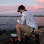 Man fishing on a rocky jetty at sunset, enjoying gear and mobile updates.