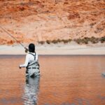 Angler fishing in tranquil lake surrounded by vibrant red rock formations and lush greenery.