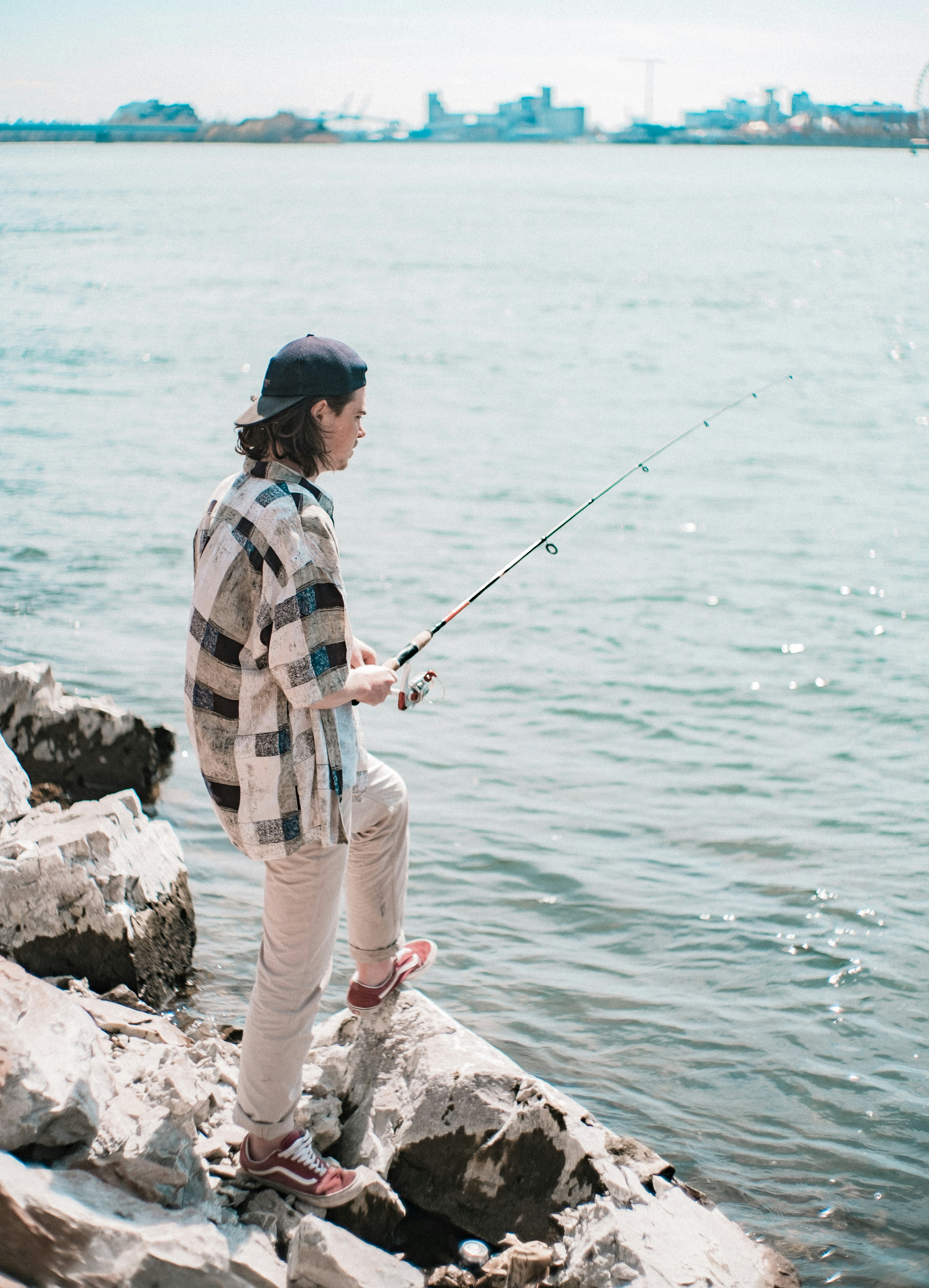 Young man fishing on rocky shoreline, showcasing casual style and urban waterfront backdrop.