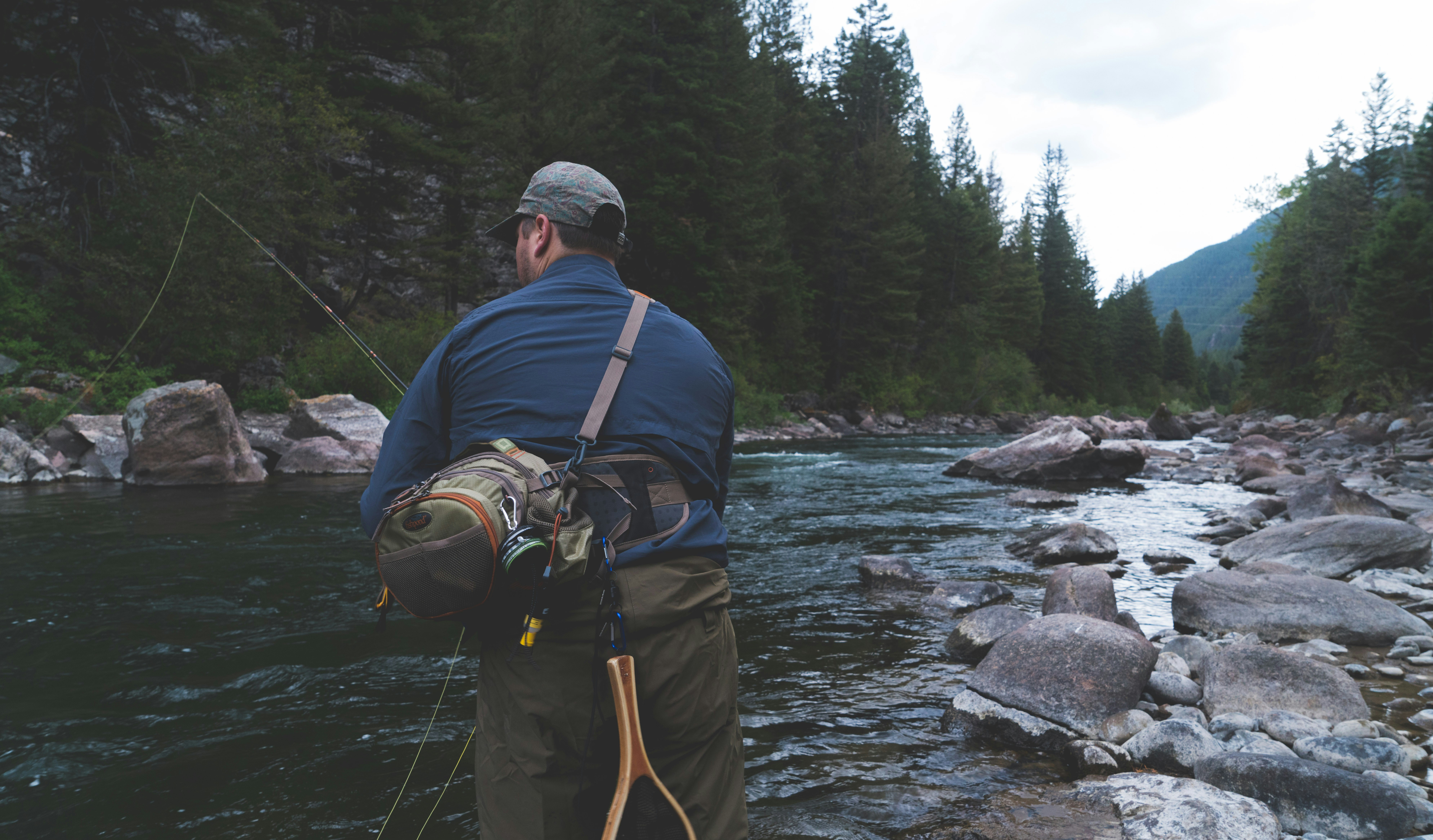 Fisherman fly fishing by a serene river in a lush, natural landscape.