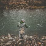 Fisherman fishing by a serene river, showcasing outdoor gear and peaceful nature scenery.