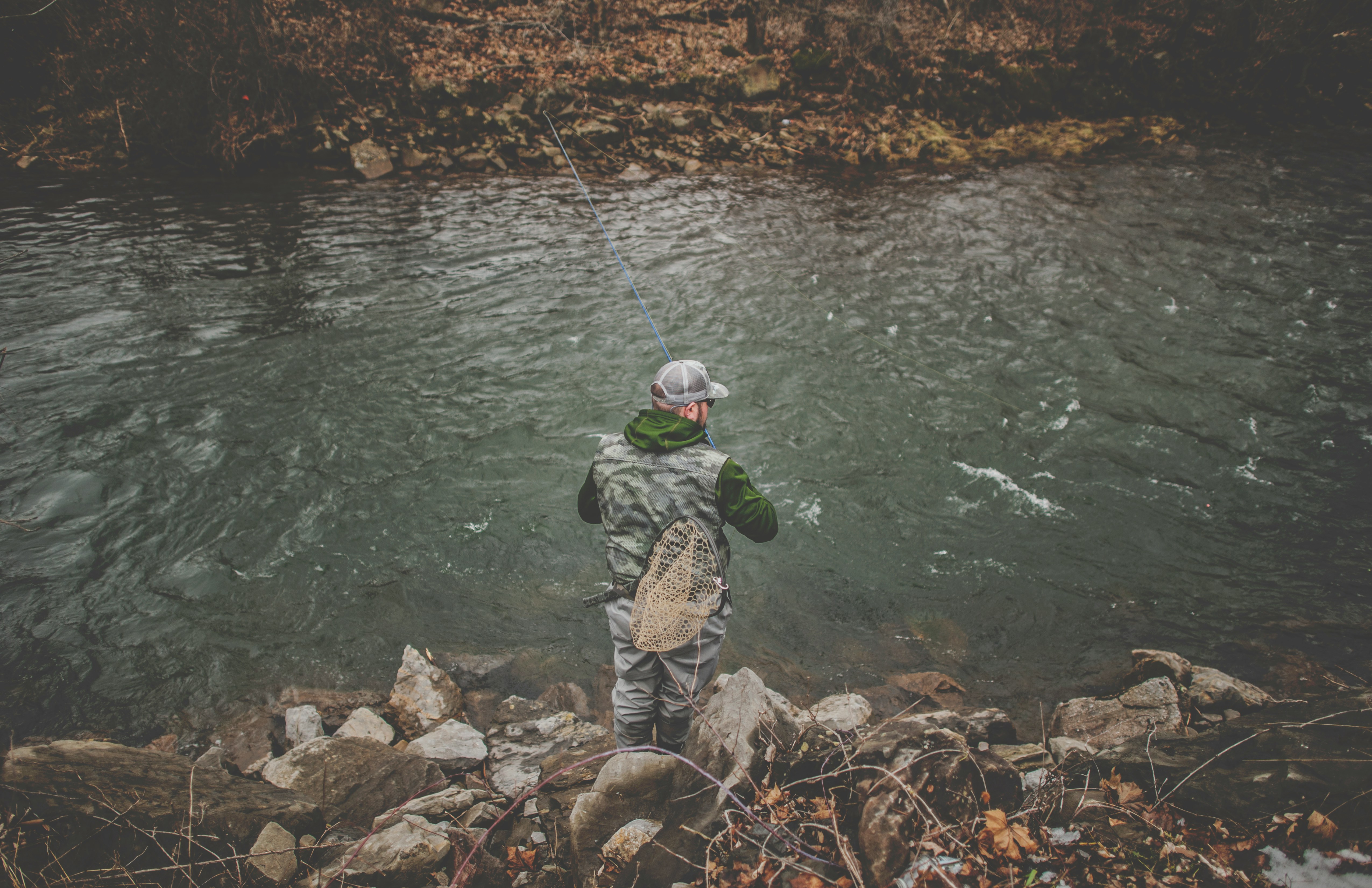 Fisherman fishing by a serene river, showcasing outdoor gear and peaceful nature scenery.