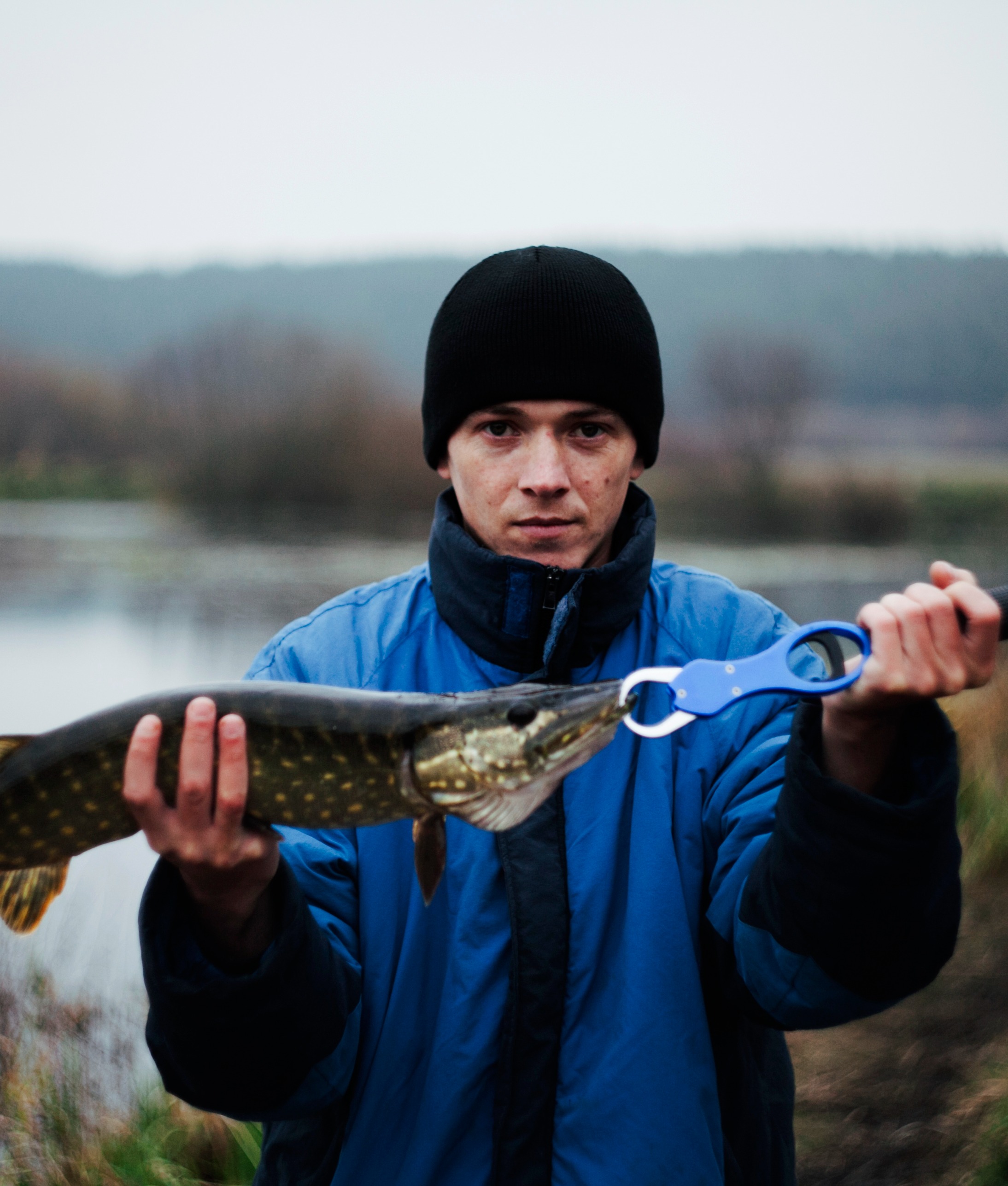 Young man proudly holds pike fish by water in serene fishing trip scene.