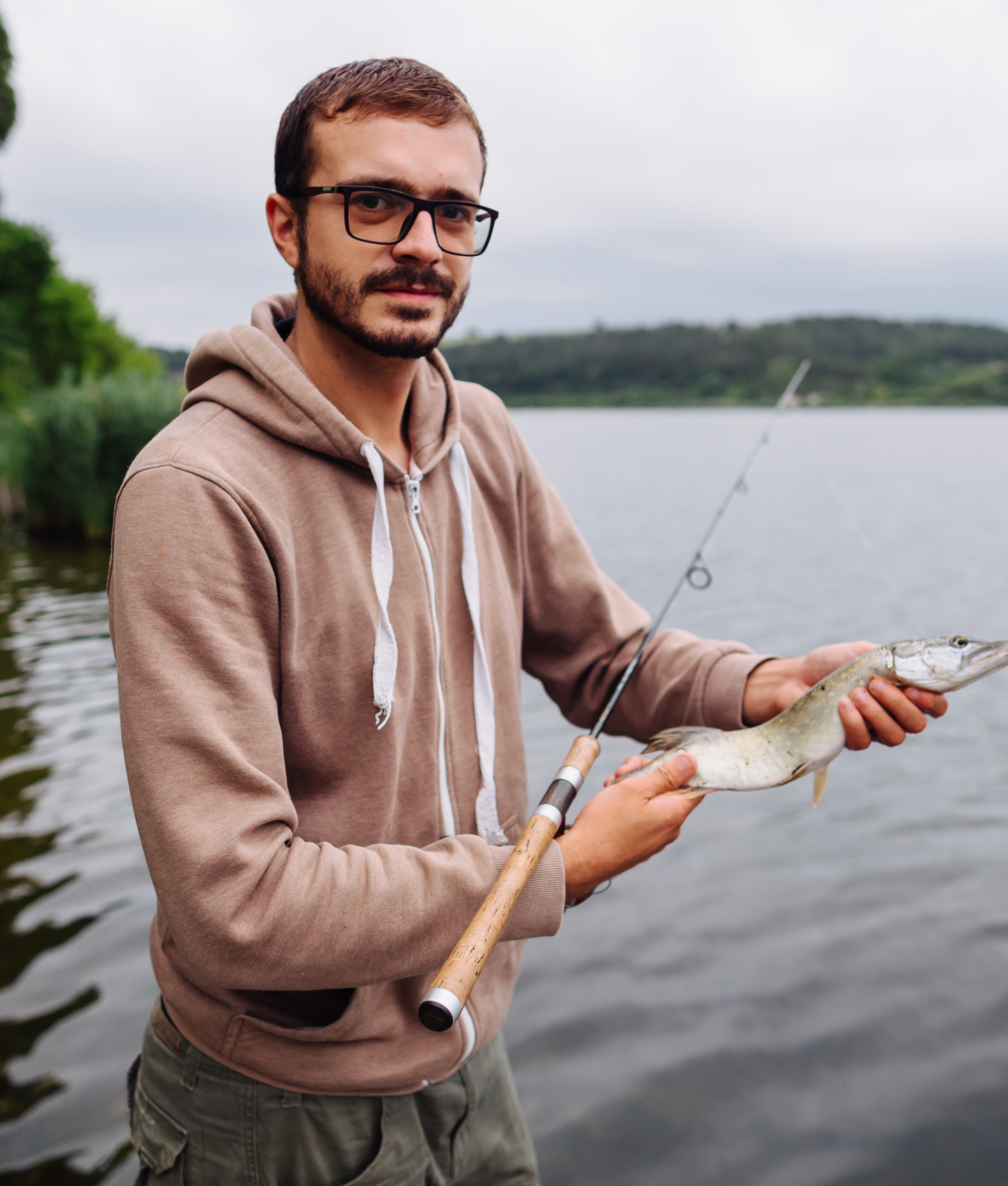 Young man proudly holds a pike by a peaceful lake during a fishing trip.