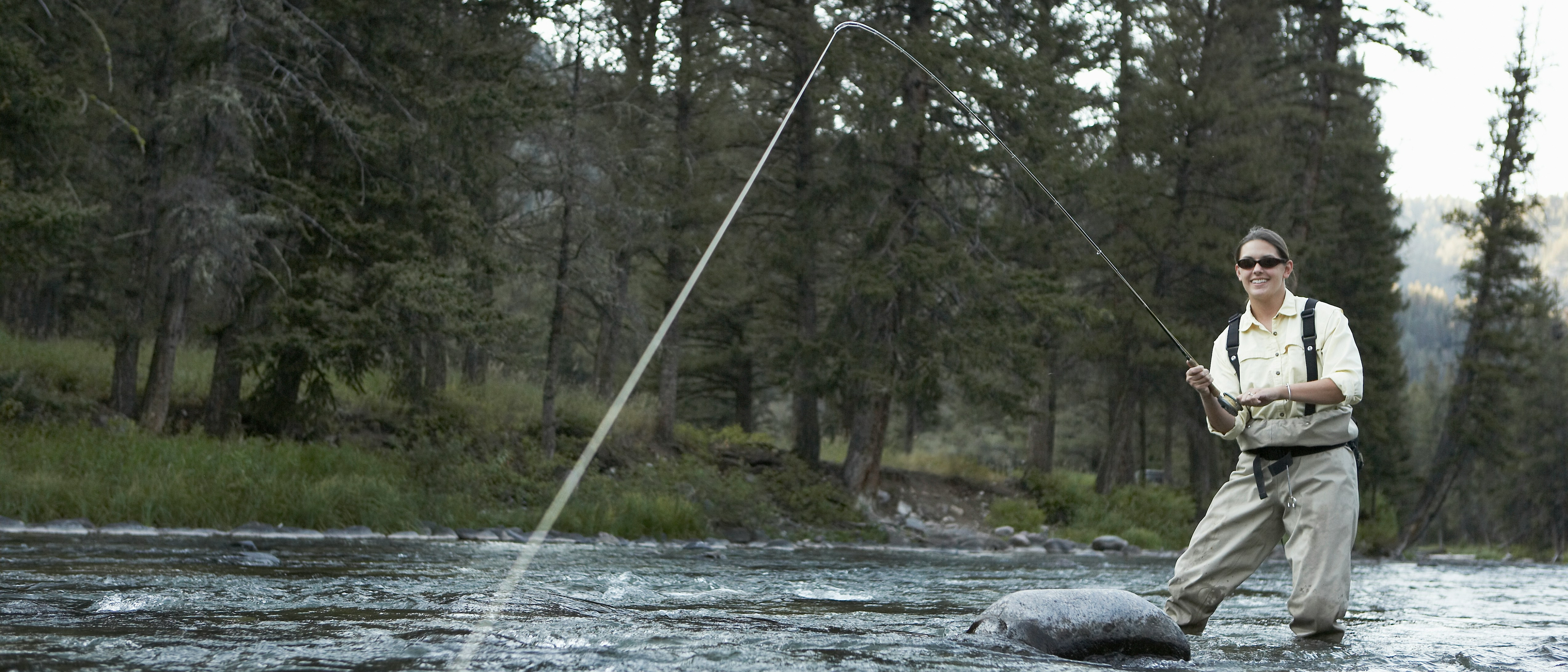 Woman fly fishing in a tranquil river, surrounded by lush nature and tall trees.