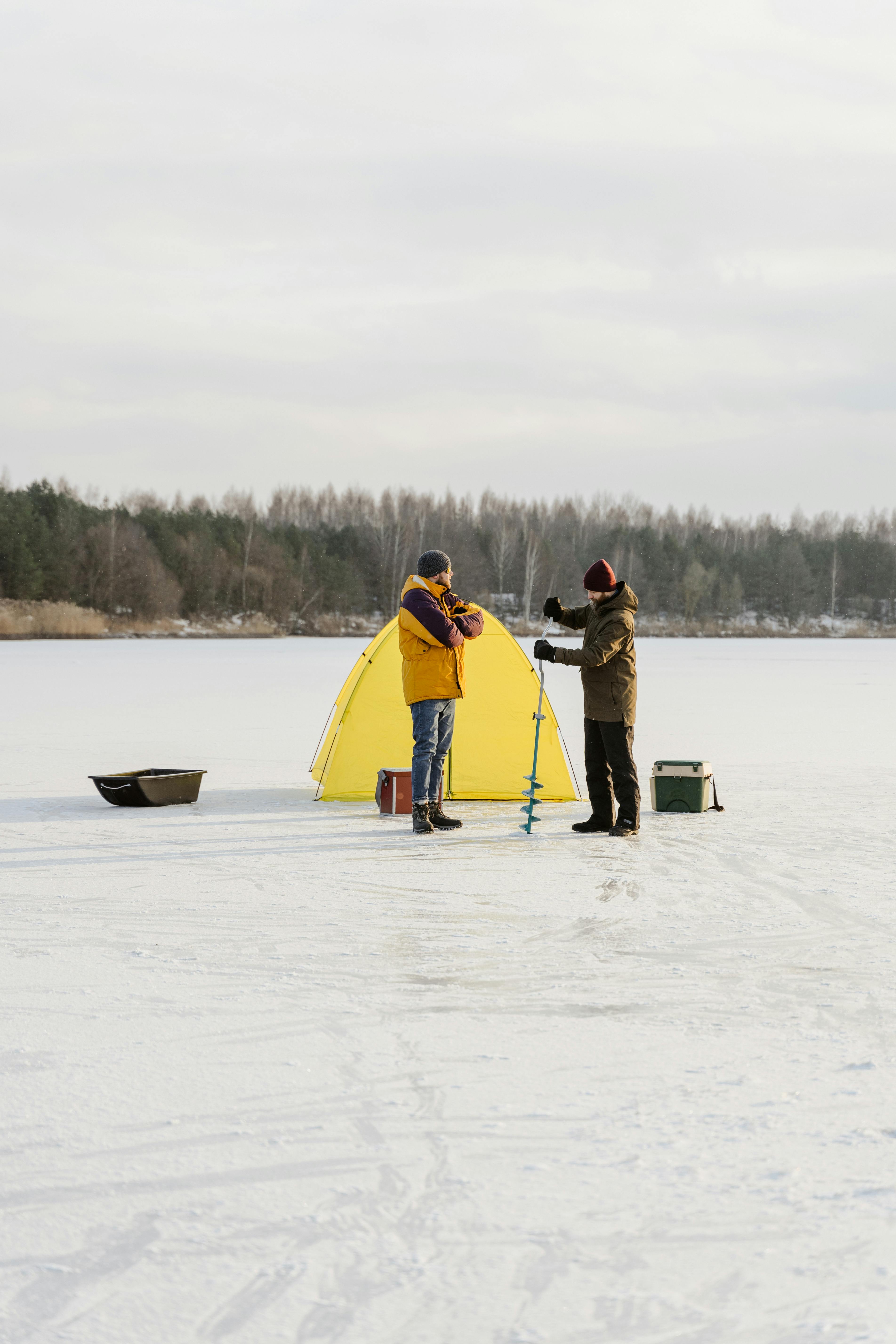 Couple enjoys ice fishing on a snowy lake, equipped with gear and a vibrant tent.