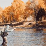 Fisherman casting a line in a serene autumn river surrounded by vibrant foliage.