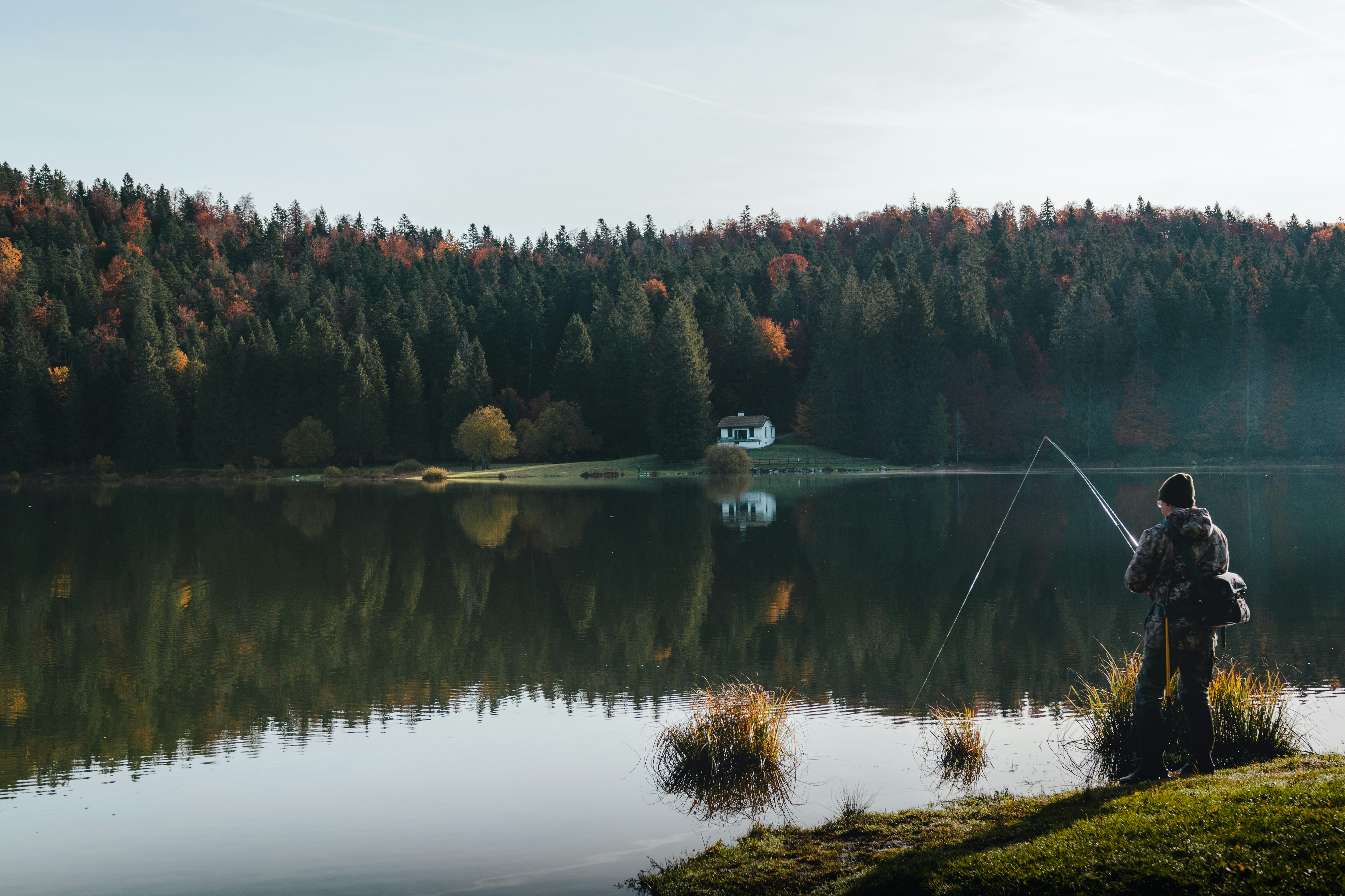 Camouflaged fisherman enjoys peaceful lakeside fishing amidst vibrant autumn colors and serene nature.