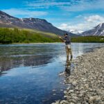 Fisherman casting a line in a serene river surrounded by majestic mountains and lush greenery.