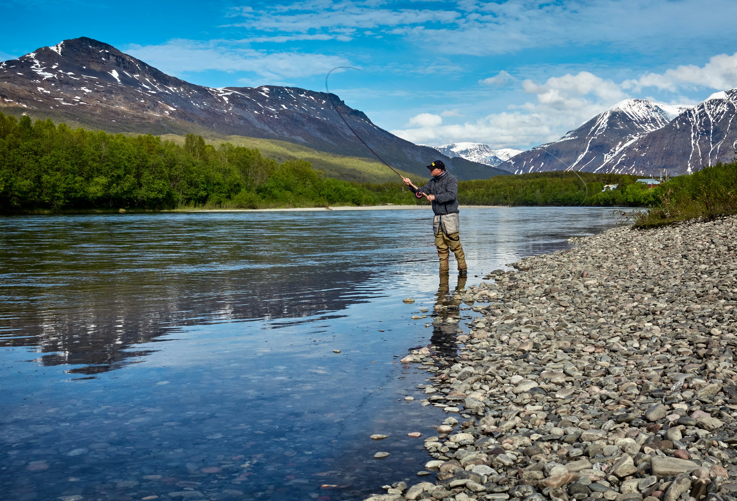 Fisherman casting a line in a serene river surrounded by majestic mountains and lush greenery.