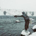 Lone angler fly fishing in a snowy winter landscape, showcasing fishing gear and calm waters.