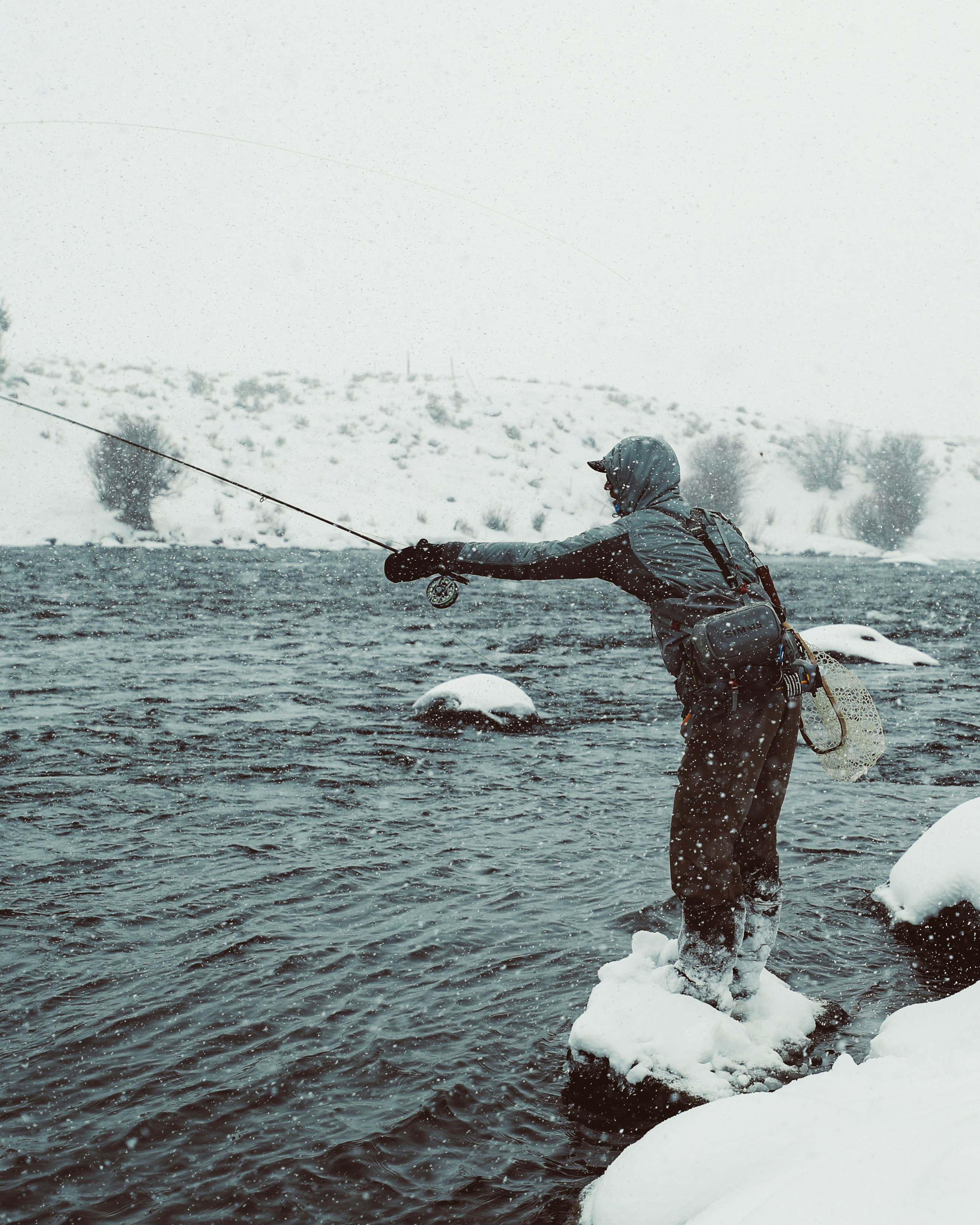 Lone angler fly fishing in a snowy winter landscape, showcasing fishing gear and calm waters.