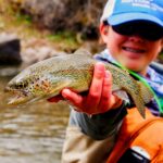 Young boy proudly holding a colorful rainbow trout during a fishing adventure.