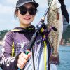 Joyful woman with stylish fishing gear proudly displays her vibrant catch by the water.