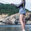 Young woman fishing on rocky shore, enjoying nature with stylish fishing gear by the water.